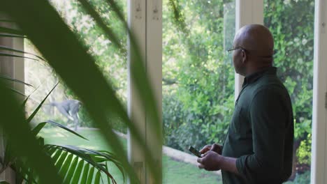 African-american-senior-man-using-smartphone-while-looking-outside-the-window-at-home