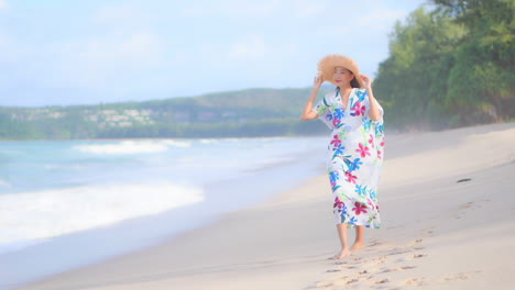 slow-motion of asian lady walking along the beach in thailand wearing a long sundress, she is holding her straw hat on windy day, slow-motion following show side view