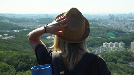 Chica-Con-Sombrero-Y-Gafas-De-Sol-Con-El-Pelo-Suelto-Apoyado-En-La-Valla-Y-Disfrutando-Del-Horizonte-Del-Centro-De-La-Montaña-Gwanaksan-Durante-El-Verano-En-Seúl,-Corea-Del-Sur