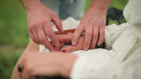 a close-up shot of a man tenderly positioning a woman's hands on her lap, highlighting her vibrant red nails and a distinctive ring, all set against the calming backdrop of a tranquil green field