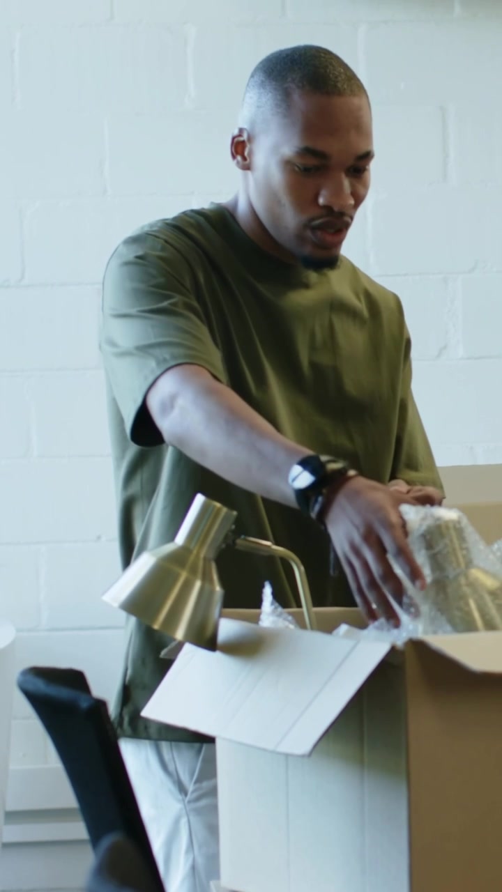 Vertical video: Lifting box to desk, man in olive tee unpacking lamp and bubble-wrap, prepping desk