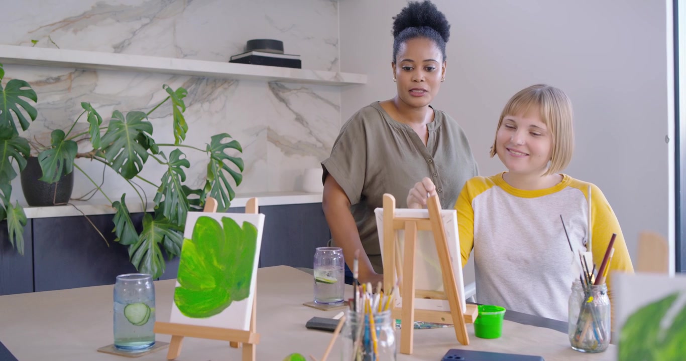 Diverse female instructor bringing water, teaching early-20s student painting on easel at studio