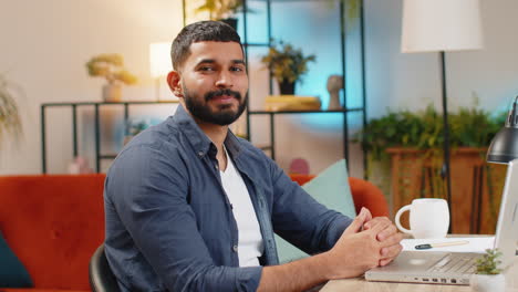 Portrait-of-smiling-Indian-man-freelancer-sitting-at-table-with-laptop-working-remotely-at-home