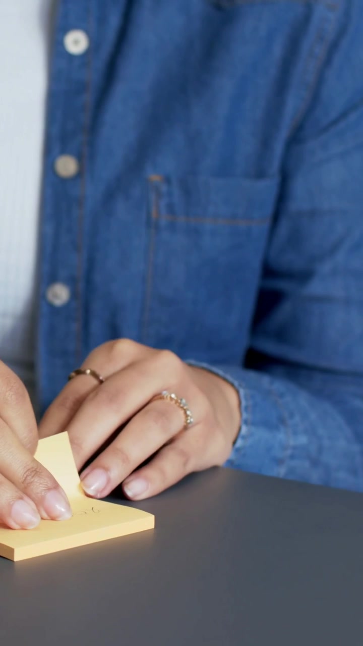 Video verticale: Una donna in jeans appoggia le mani, leviga e stacca una nota gialla vicino al laptop per riferimento.