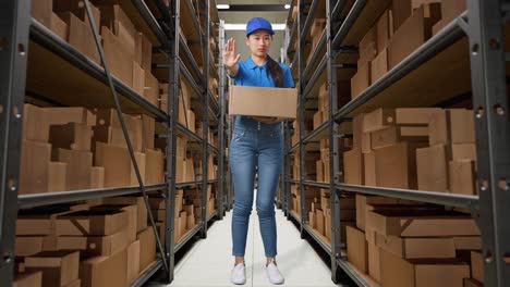 full body of asian female courier in blue uniform making stop gesture and shaking her head while delivering a carton in warehouse