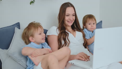 Young-beautiful-mother-and-two-young-children-sitting-on-the-couch-looking-at-the-laptop-screen-and-make-online-purchases