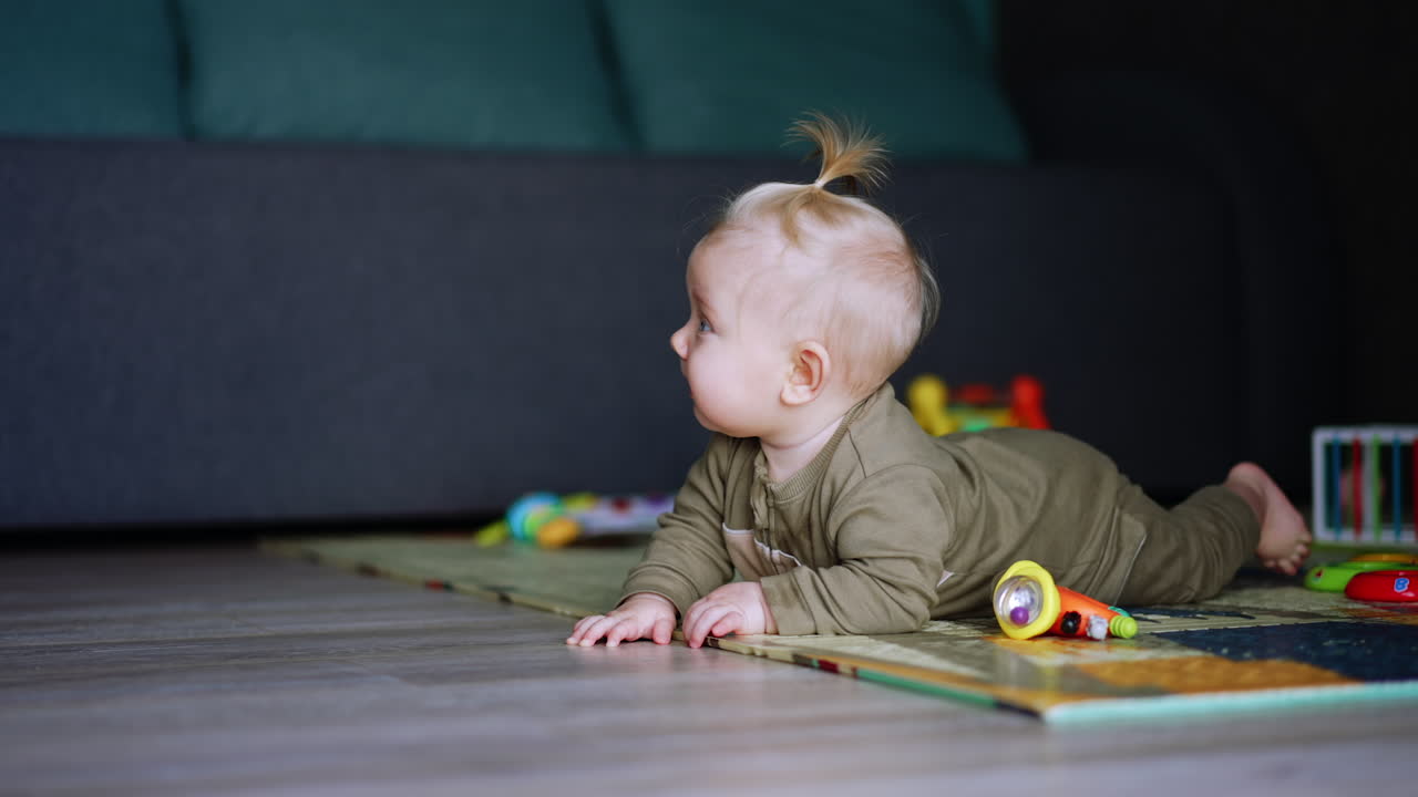 Lovely baby in bodysuit lies on the floor. Side view of a cute kid with little pony tail on his blond hair.