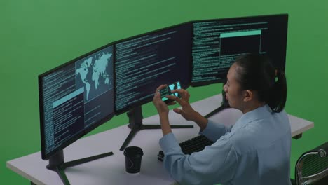 side view of asian woman developer working with mobile phone while write code with multiple computer screens in green screen studio