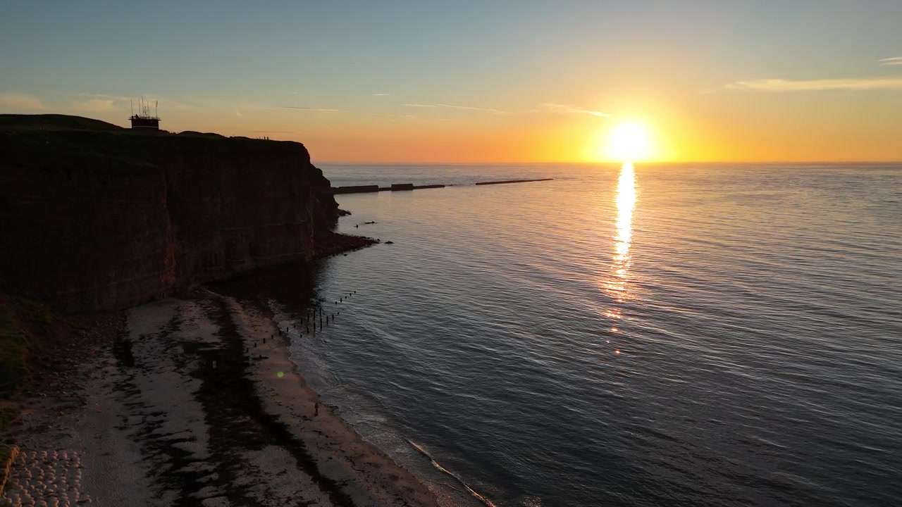 Drone vetäytyy taaksepäin auringonlaskun siluetiksi rannan meren ja tummien Helgolandin kallioiden ylle.