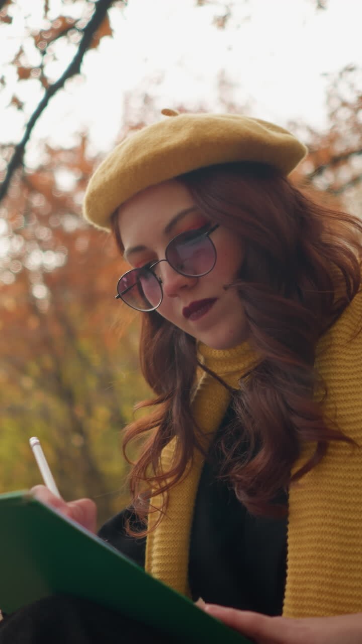 Vista laterale di una signora seduta che disegna nel suo quaderno, profondamente concentrata, sullo sfondo di alberi autunnali e foglie colorate in un ambiente esterno tranquillo.