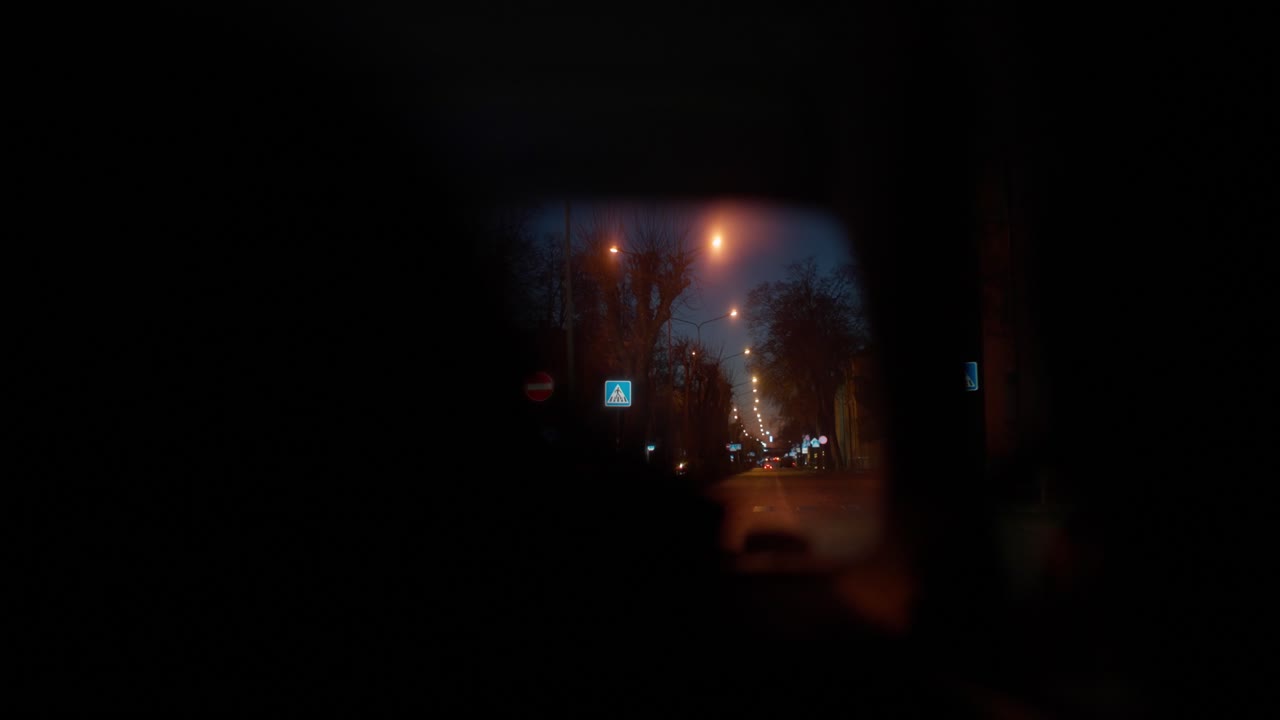 Inquadratura POV dal sedile posteriore di un'auto che guida per le strade della città al crepuscolo. Luci stradali e cartelli stradali visibili attraverso la stretta apertura del finestrino nell'oscurità