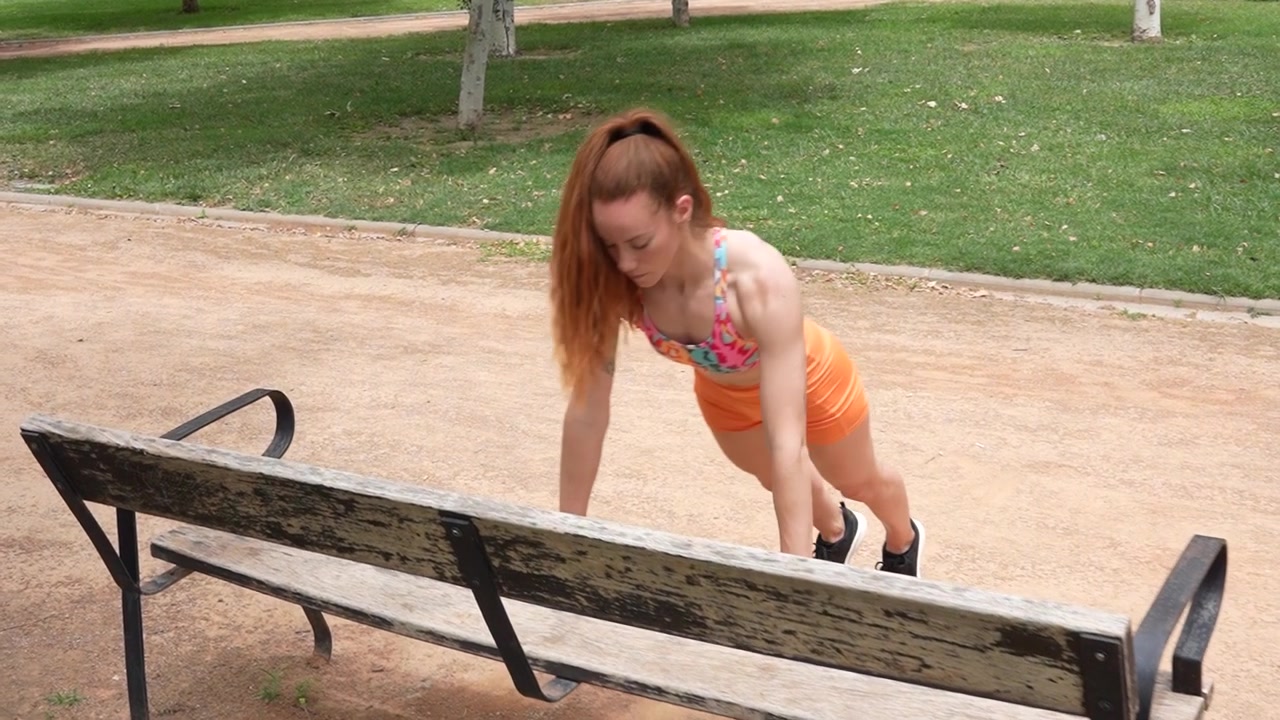 Una donna sta facendo flessioni inclinate su una panchina in un parco verde, allenandosi con determinazione.