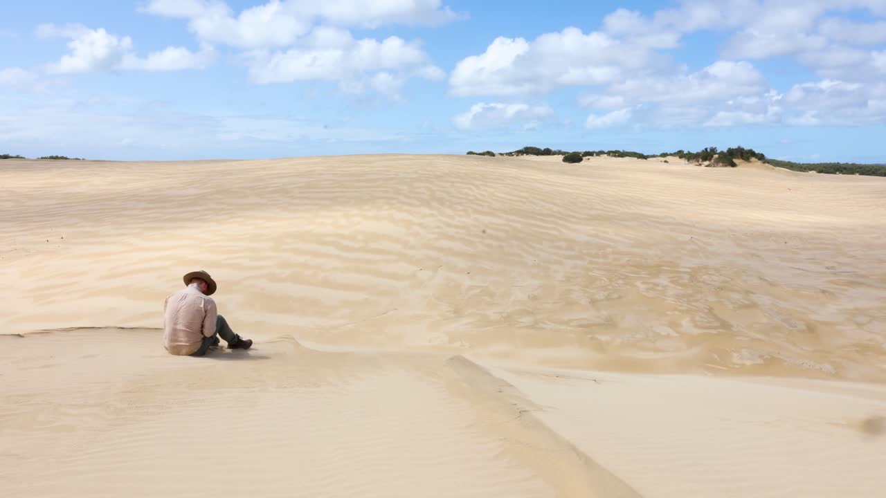 아쿠브라를 입은 호주 부시맨이 장엄한 모래언덕 가장자리에 앉아 있습니다.