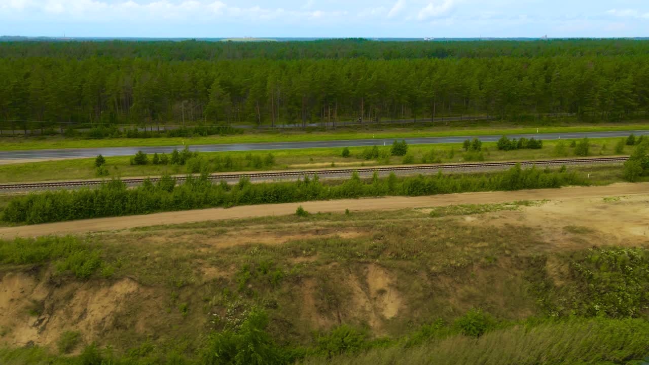 Aerial drone footage flying besides a rural countryside highway where cars and vehicles drive next to a open pit sand quarry mine where large blue lake is visible in front and forest in the horizon