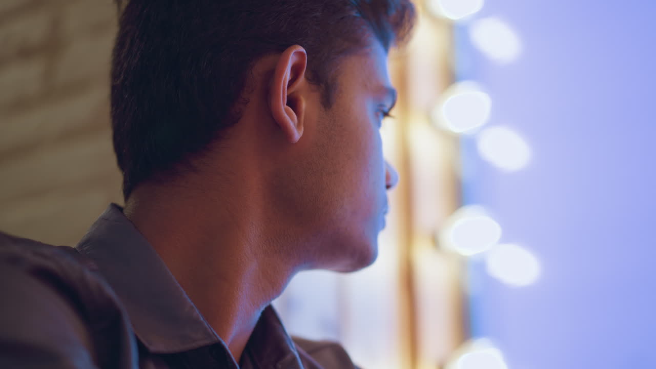 Rear view of young man sitting indoors facing softly lit blue window, head slightly turned in quiet contemplation, surrounded by gentle ambient glow, suggesting mood of solitude, thoughtfulness, and relaxation