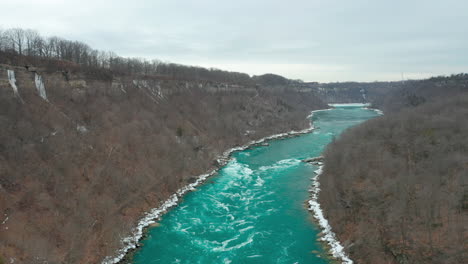 Aerial-shot-of-river-flowing-through-dead-forest-at-winter-of-fall-time-under-cloudy-skies