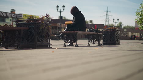 back view of white girl seated in public space, focused on her book, with flower pot swaying gently in wind and pedestrians walking in background, stores with inscription visible across the road