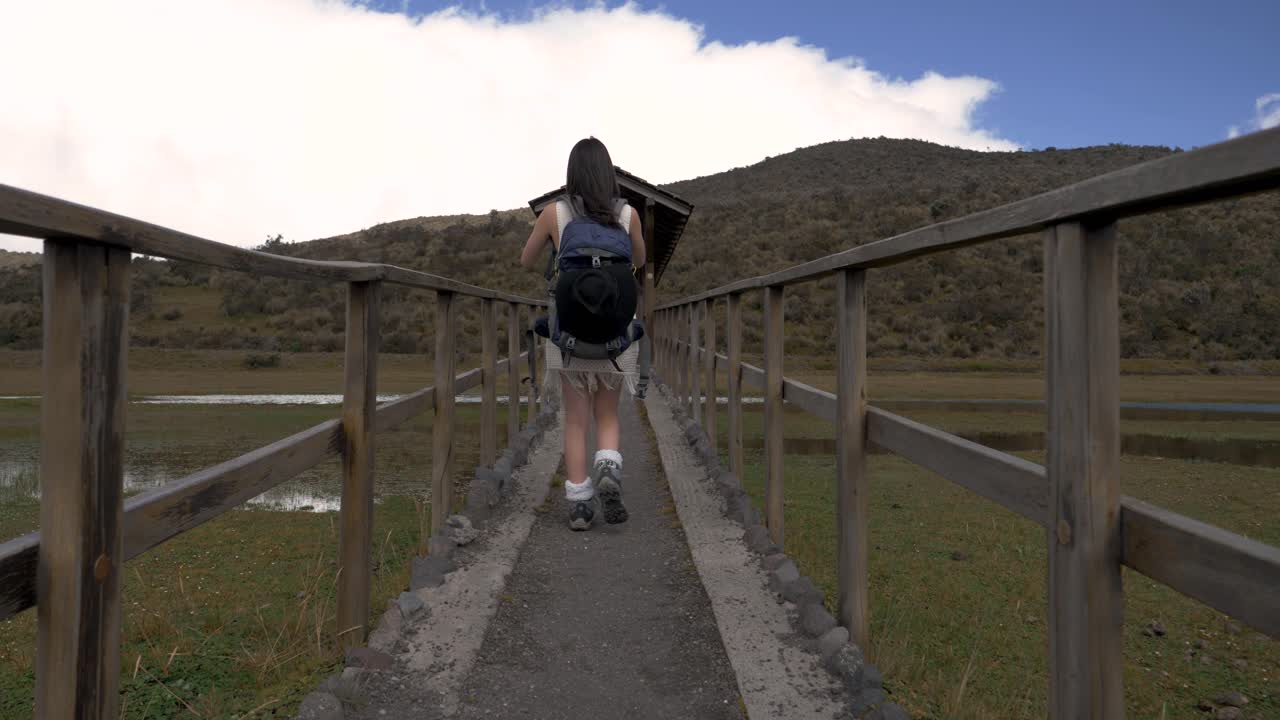 Rear tracking follow woman with hiking backpack walk across wooden bridge in cotopaxi national park