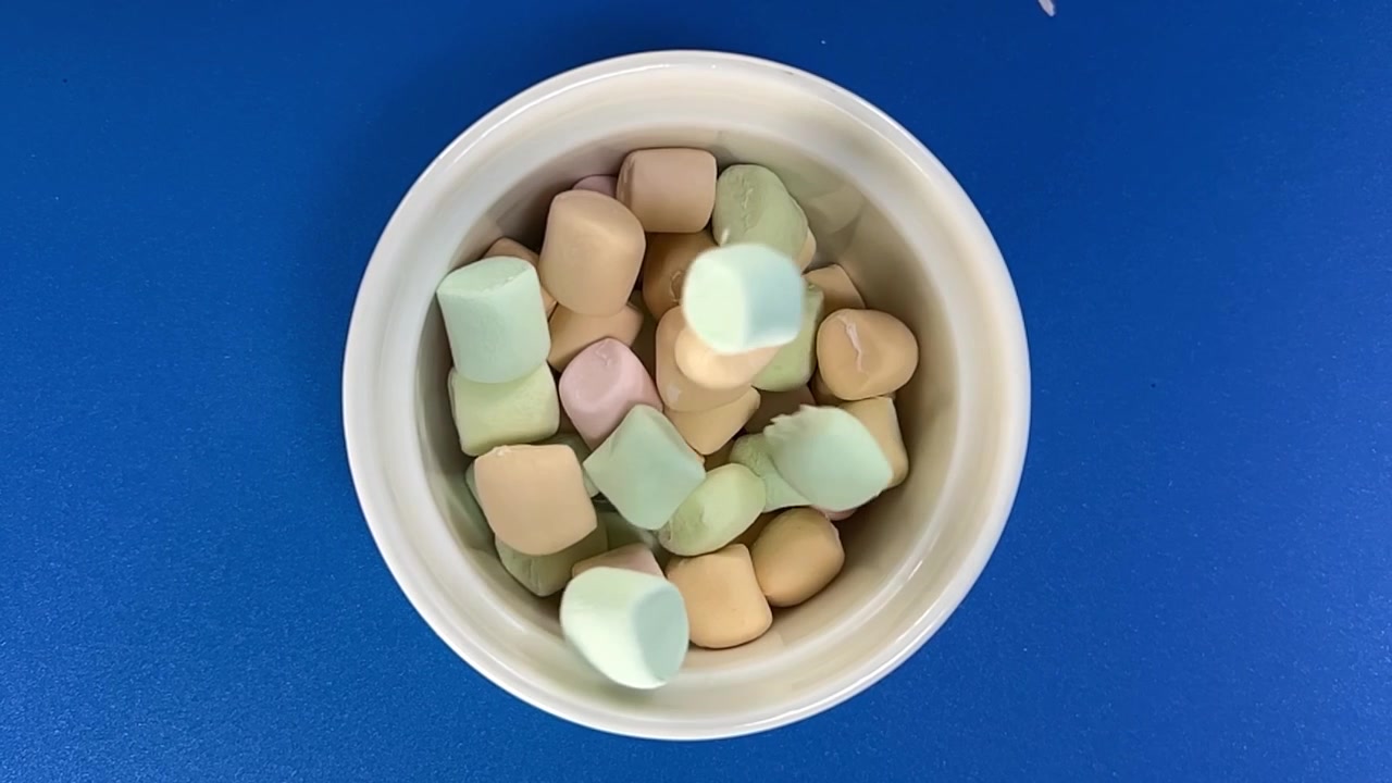 Vista dall'alto di marshmallow colorati che cadono in una ciotola bianca su uno sfondo blu, catturando pezzi di caramelle morbide in movimento con una composizione alimentare giocosa
