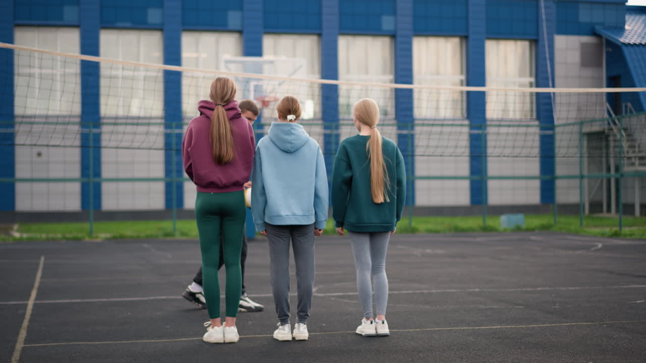 볼리볼 코트에 서 있는 스포츠 의상을 입은 젊은 운동 선수들의 뒷면, 코치가 공을 는 것을 지켜보고, 그들의 머리카락이 바람에 날아다니고, 네트가 있는 야외 코트, 흐릿한 배경의 농구 코트