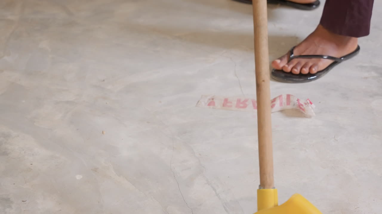 Esfregão rosa varrendo piso de concreto da casa com cabo amarelo, sandálias pisando por perto, poeira e resíduos acumulados, movimento de limpeza casual e varredura rítmica sugeridos, dona de casa realizando tarefas diárias.