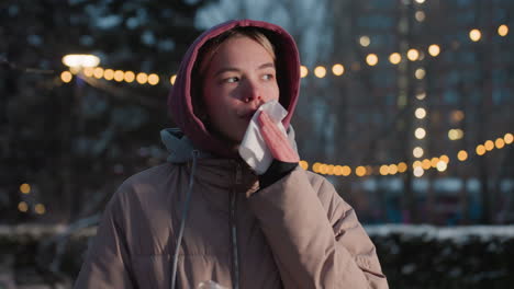 dama sosteniendo un bocadillo en una mano mientras usa una servilleta para limpiar la boca en un entorno al aire libre de invierno con luces bokeh en el fondo, vestida con una sudadera con capucha y chaqueta cálida