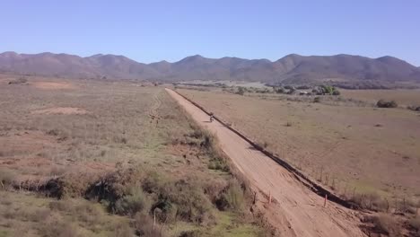 Conducción-De-Motocicletas-Sola-En-Camino-Rural,-Montaña-En-El-Horizonte,-Seguimiento-Aéreo