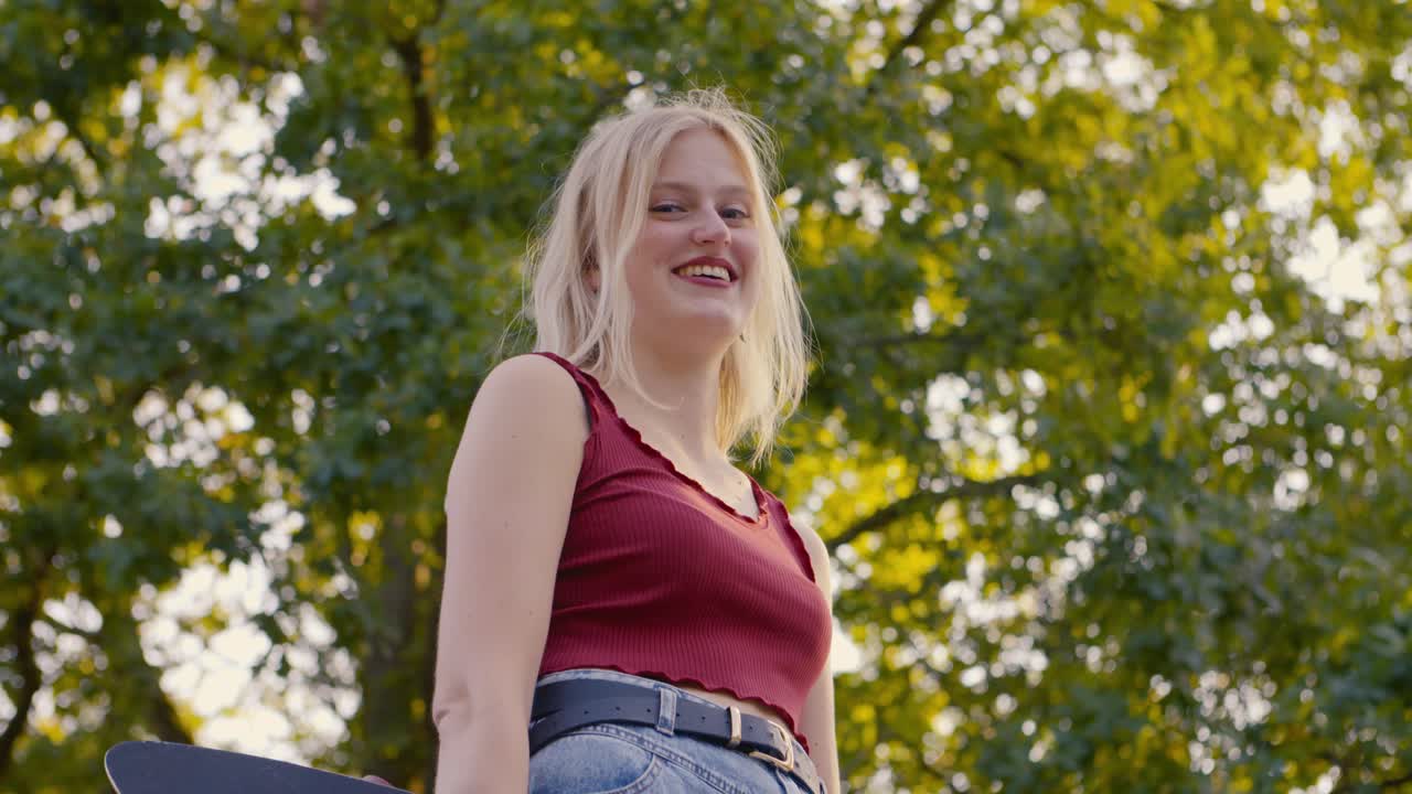 카메라가 천천히 올라가는 동안 웃으면서 유혹하는 스케이트보드를 들고 있는 소녀는 스케이트 공원에서 고해상도로 카메라를 보고 웃는다