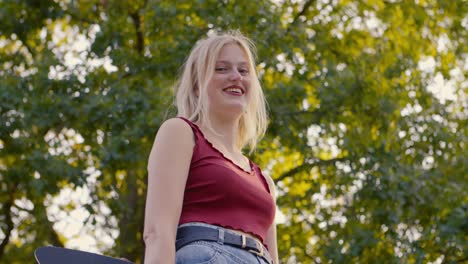 girl holding a skateboard posing laughing and flirting while the camera slowly goes up and she smiles at the camera in a skate park, in high resolution