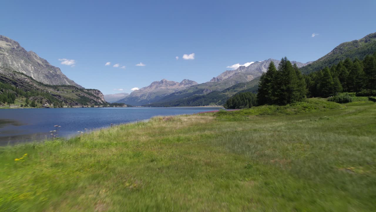 Flyfoto i kinostil av en grønn eng ved siden av en elv og noen fjell i horisonten i Silvaplana, Sveits