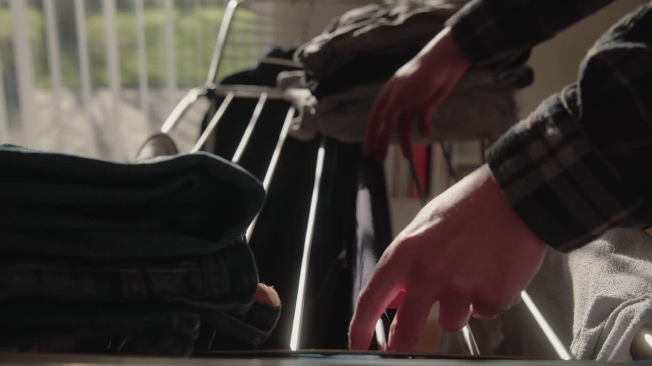 Young Male collecting and folding dry clean Clothes from Dryer At Home in sunlit modern living room Black T-Shirt Detail 4K