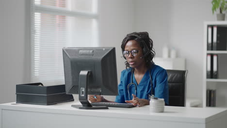 una mujer negra que usa auriculares se sienta en una mesa con una computadora y recibe llamadas de pacientes mira sus registros médicos y los ingresa en el horario de la clínica