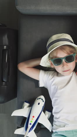 relaxed preschooler adjusts stylish sunglasses lying on sofa near suitcase and plane toy in yacht cabin. cute girl spends vacation on seaside upper close view