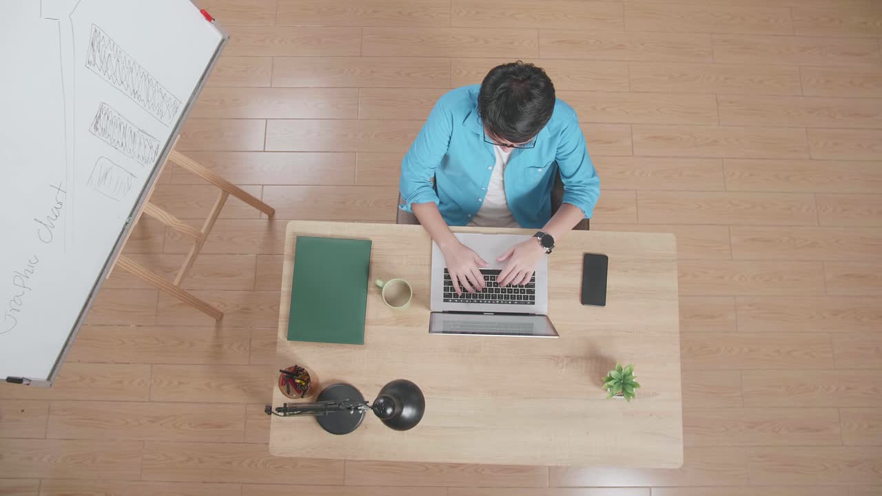 뷰  아웃  한 남자가 집에 있는 화이트보드 에 있는 나무 책상에 앉아서 스마트폰 에 노트북으로 타이핑하고 있다.