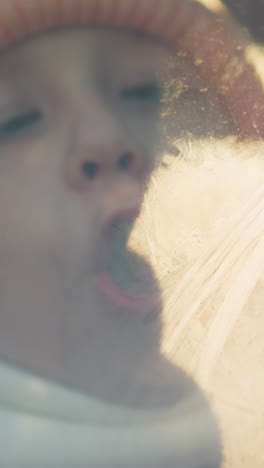 a close-up of a young girl wearing a pink knit hat and white scarf, blowing air onto a foggy car window. her breath creates condensation on the glass