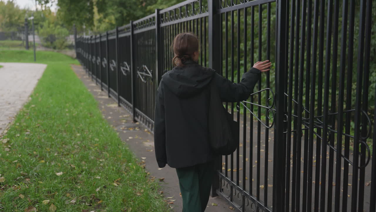 persona caminando junto a una valla de hierro bajo un cielo nublado, la silueta de una chaqueta oscura se mueve junto a una barandilla negra alta que bordea un sendero frondoso del parque, la mano roza los barrotes, ritmo lento y reflexivo, hojas caídas sobre la hierba