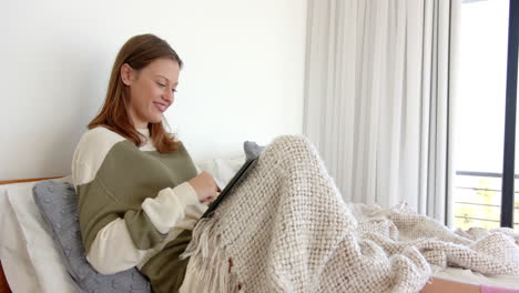 relaxing on bed, young woman using tablet and smiling under cozy blanket