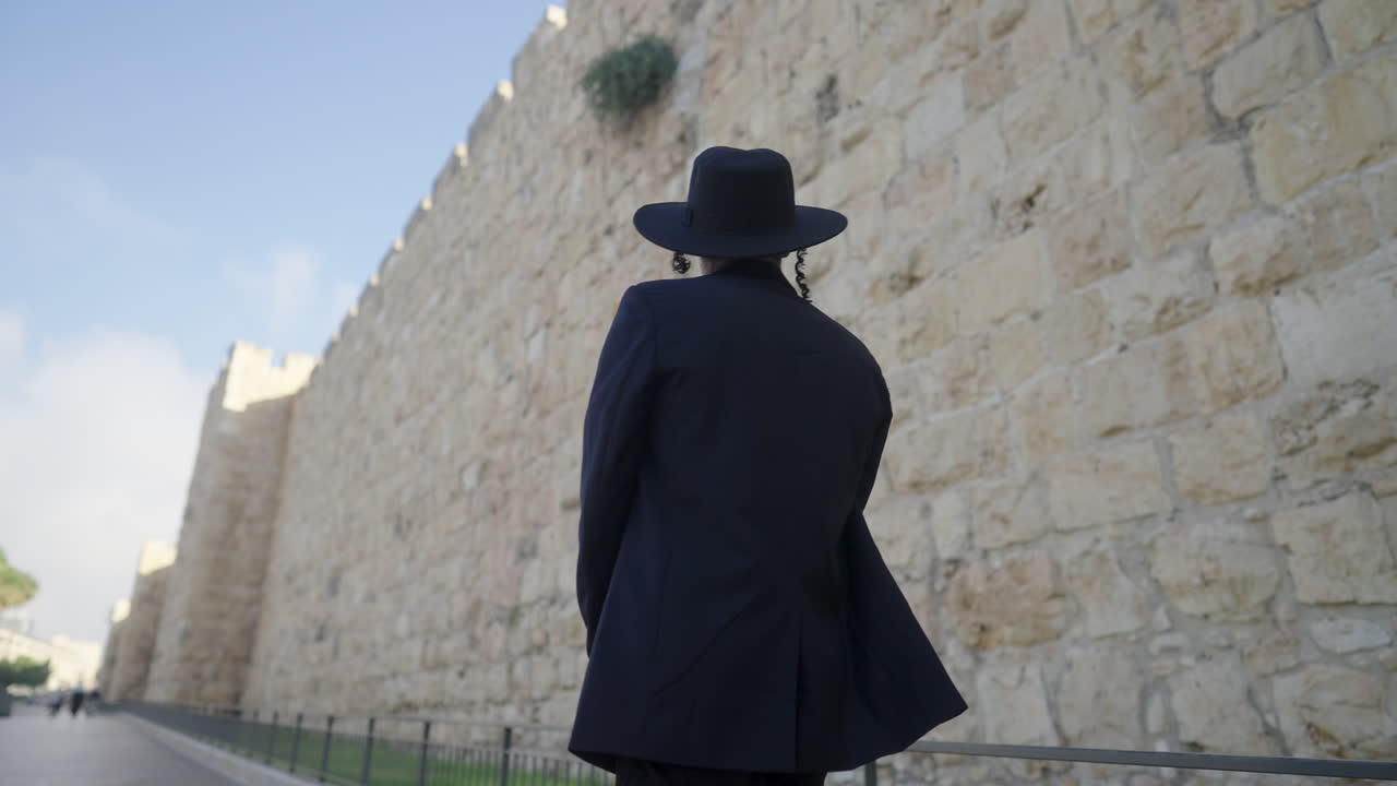 Rear view of an Orthodox male in a black suit and hat, walking along a pathway, gazing at the ancient architecture of the Old City walls in Jerusalem