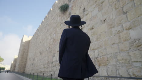 rear view of an orthodox male in a black suit and hat, walking along a pathway, gazing at the ancient architecture of the old city walls in jerusalem