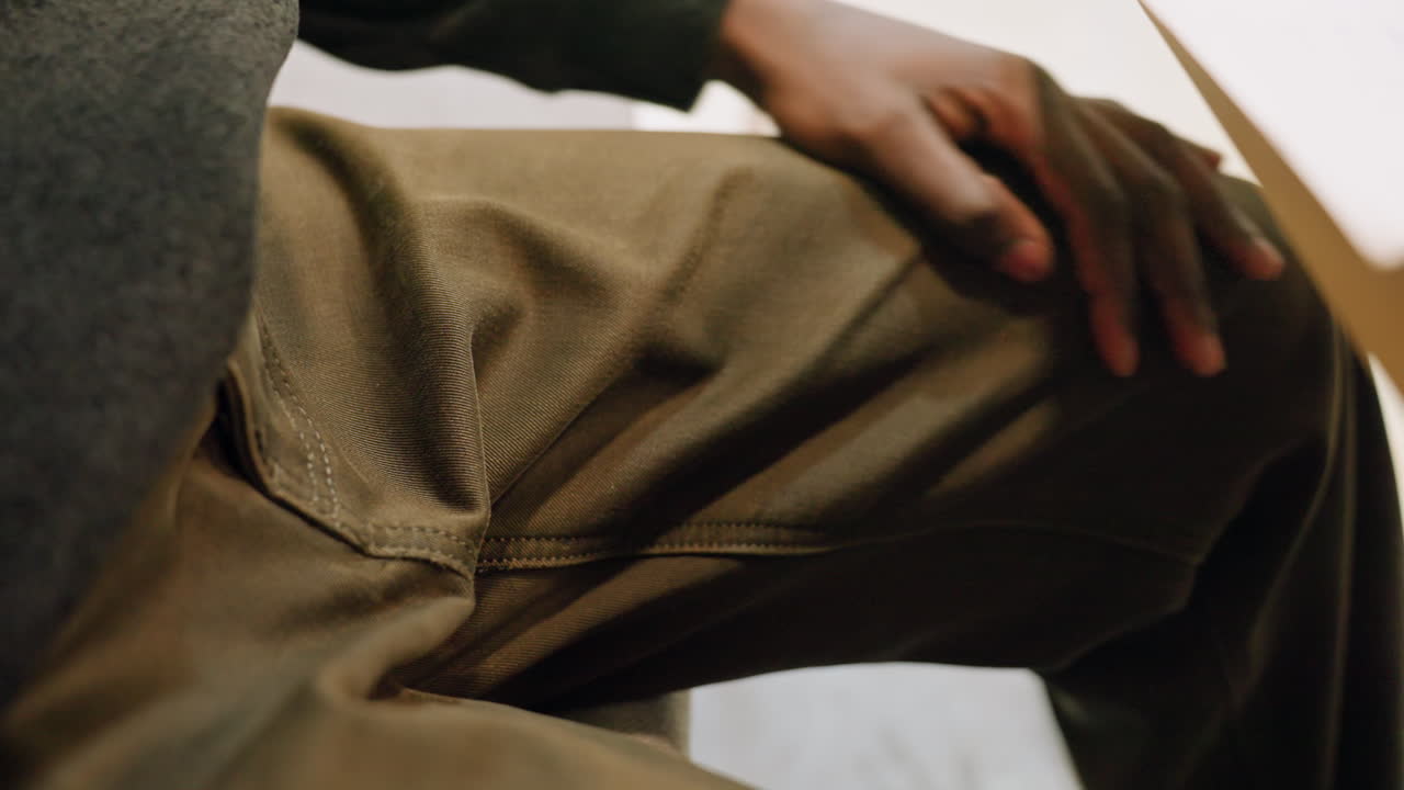 Hand on thigh black man, olive pants and gray sweater, subtle tapping and fidgeting, nervous candidate during interview or waiting room, shifting posture, desk edge, quiet tension, close cropped frame