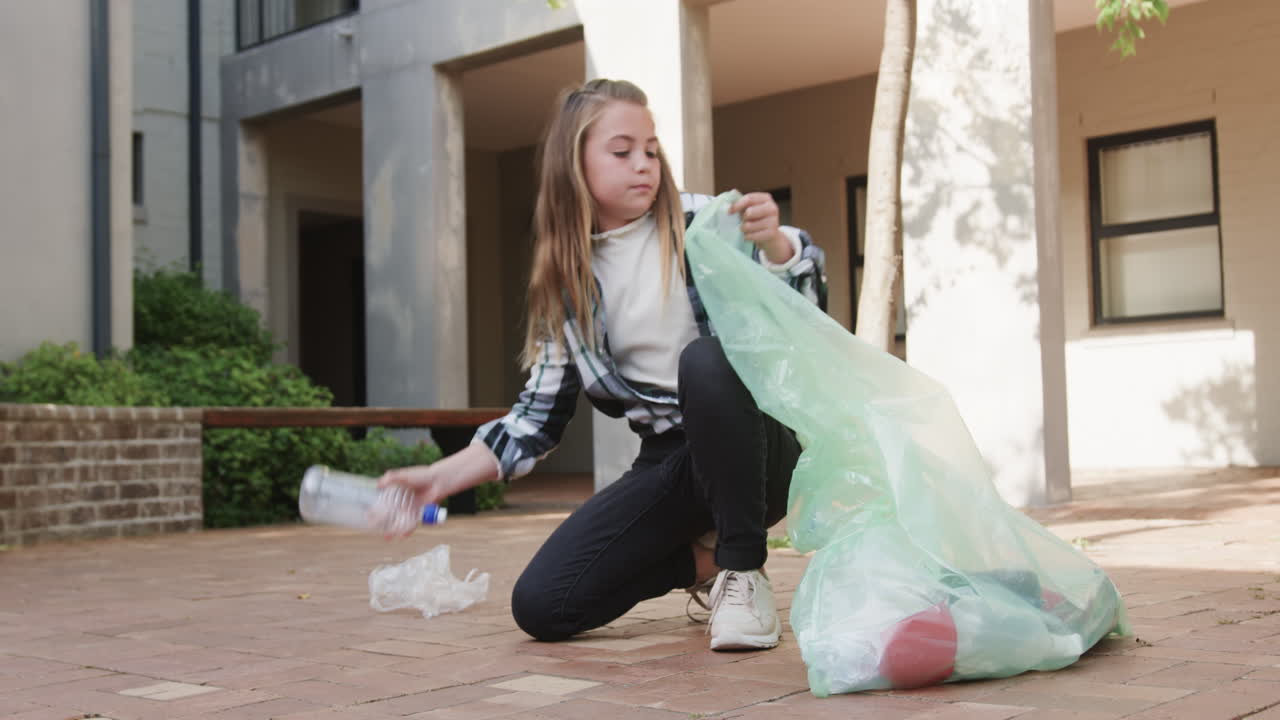 Young girl cleaning up courtyard, holding large trash bag, focused on task, at school