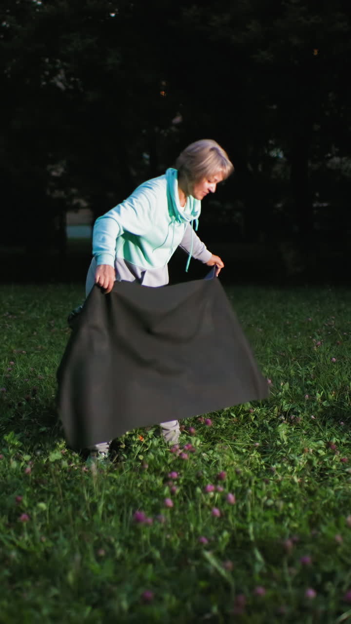 Una mujer mayor extiende una manta oscura en un parque cubierto de hierba al atardecer, con tréboles bajo sus pies y un beagle olisqueando cerca. La mujer se arrodilla para alisar la tela, vestida con un suéter informal y un bolso en la cadera, disfrutando de la tranquila rutina de una compañía al aire libre.