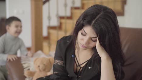 portrait of a tired middle eastern mother sitting on the armchair and her two little children jumping at the background