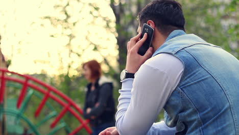 Young-guy-using-phone-on-playground-with-children-playing-in-outdoor