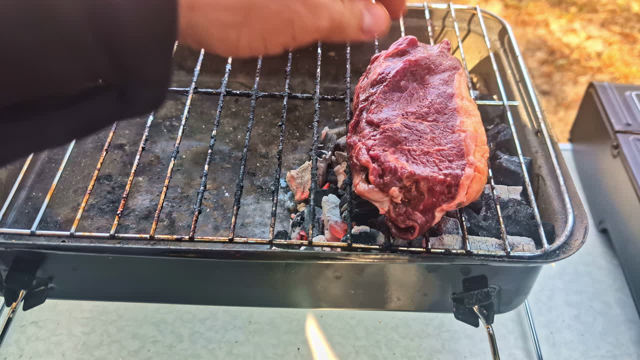 Hand placing raw marbled steak onto small charcoal grill at outdoor picnic setup