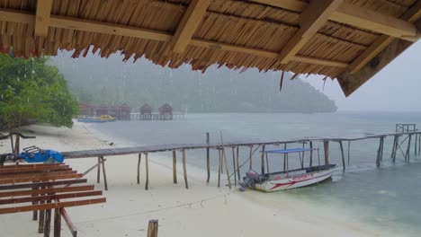 Der-Tropische-Regenschauer-Auf-Der-Insel-Kri-Im-Raja-Ampat-Archipel,-Der-Auf-Das-Meer-Und-Den-Sandstrand-Herabfällt,-Gesehen-Vom-Schutz-Eines-Holzhauses-Aus