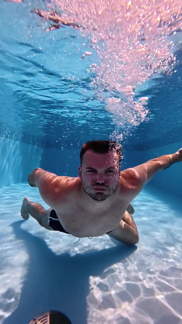 Uomo che espira aria sott'acqua mentre nuota in una piscina blu trasparente, creando un flusso di bolle intorno al suo viso in super slow motion a 240 fps