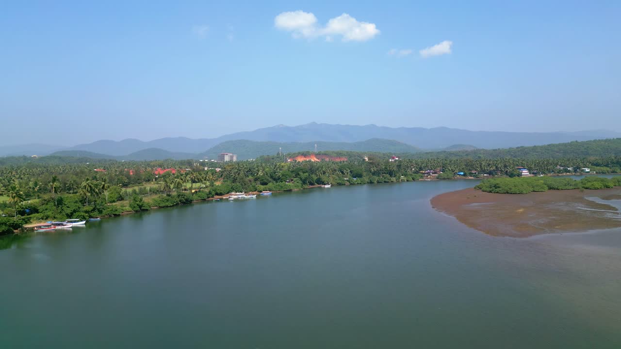 Aerial view of serene Talpona River near Rajbag Beach in Goa sparkles under the sunny sky, flanked by lush green trees and distant mountains, creating a peaceful, picturesque tropical landscape.