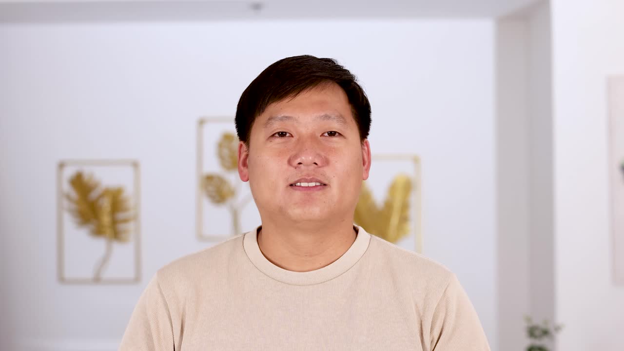 An Asian man stands indoors, hands pressed together in a respectful greeting gesture, with soft lighting and a neutral, modern background. Camera remains steady