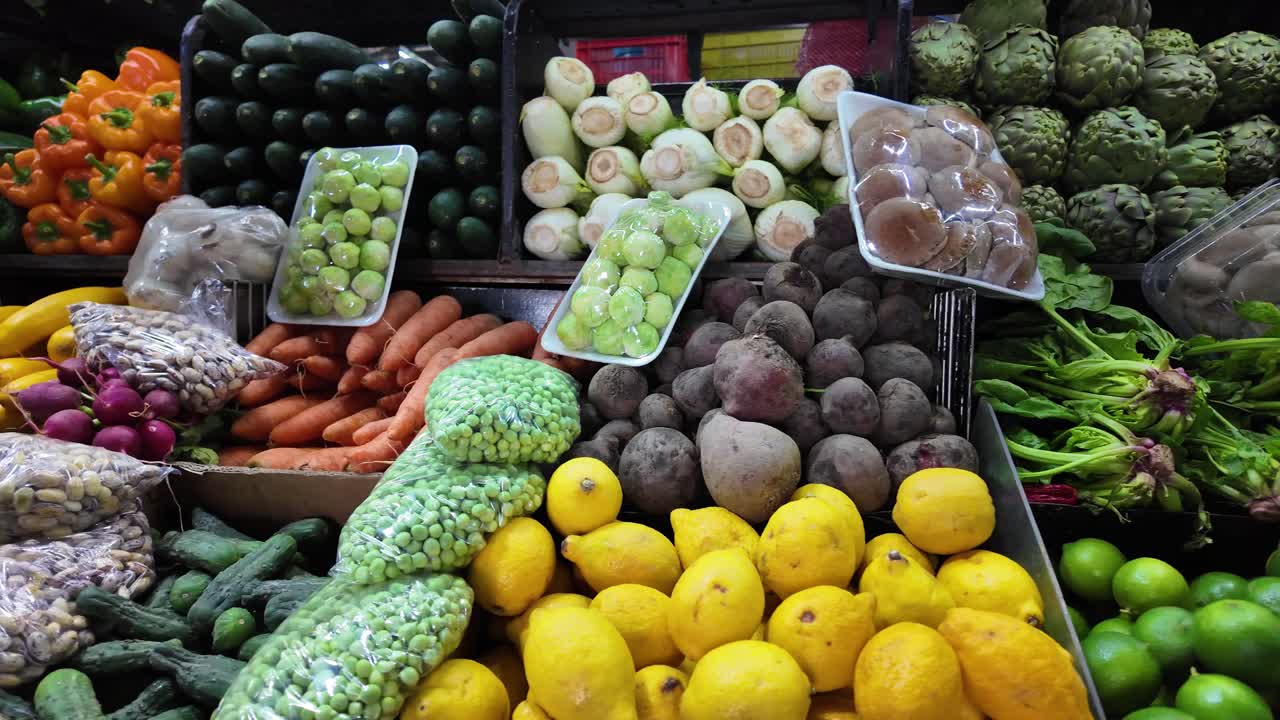 Travelling shot de uma prateleira de mercado exibindo limas coloridas, alcachofras, cogumelos, repolho, beterrabas e rabanetes brancos em um mercado agrícola